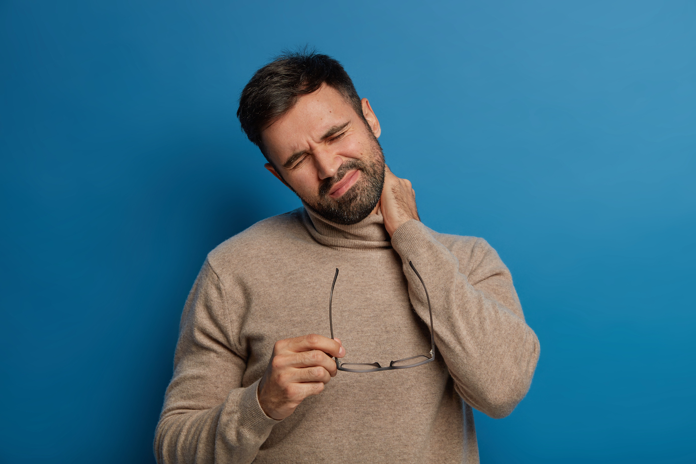 Fatigue displeased bearded man feels discomfort in neck, has problems with spine, takes off spectales, frowns face from pain, suffers without painkillers, being unhealthy and tired, isolated on blue